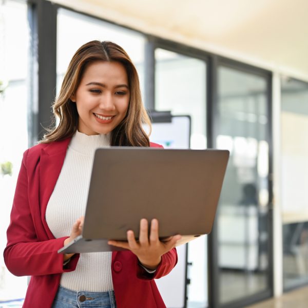 Professional and charming millennial Asian businesswoman stands in her office using portable laptop computer.