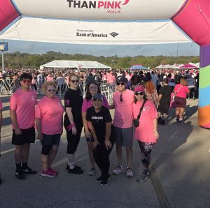Group wearing pink shirts at a charity walk event under an archway, surrounded by a large crowd.