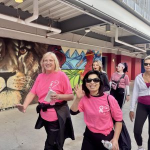 Women in pink shirts walking and smiling in front of a mural with a lion and colorful designs.