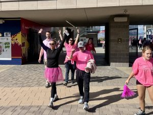 People in pink walking in an outdoor event, some raising arms, wearing shirts and tutus, one holding a pom-pom.