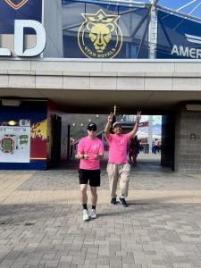 Two men in pink shirts pose outside Utah Royals stadium entrance.