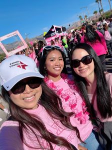 Three women smiling at a breast cancer awareness event, wearing pink outfits and sunglasses.