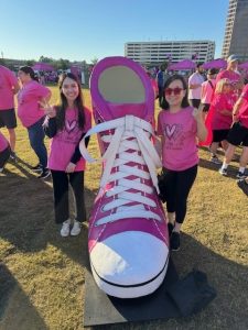 Two people in pink shirts pose with a large pink shoe at an outdoor event.