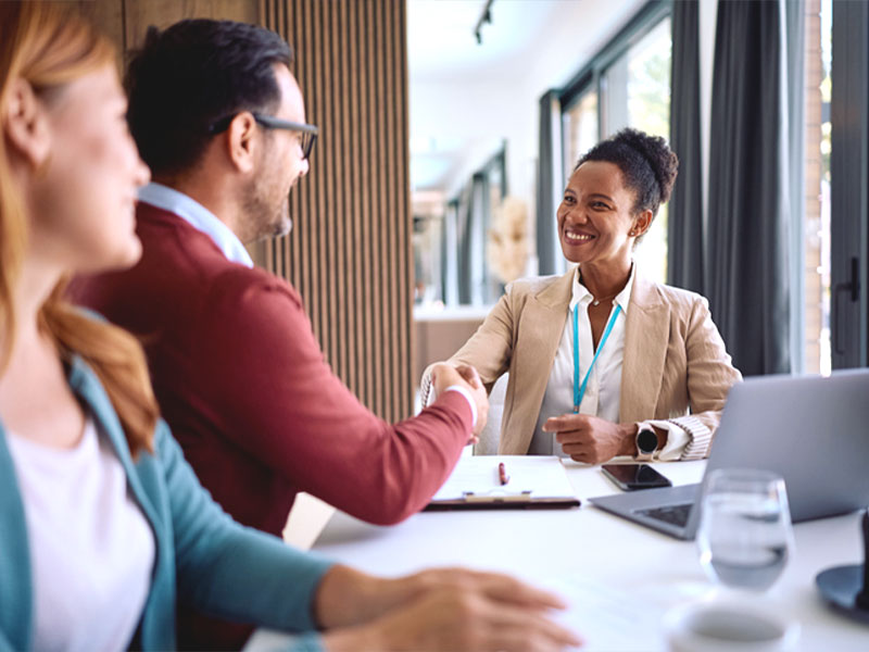 A woman in professional attire shakes hands with a man across a table, smiling during a business meeting.