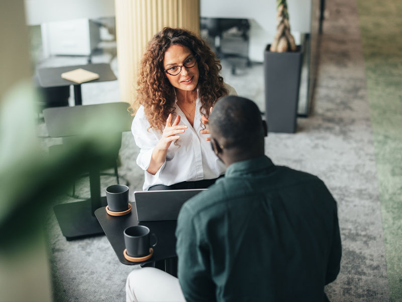 Two people having a discussion at a table with coffee mugs and a laptop.