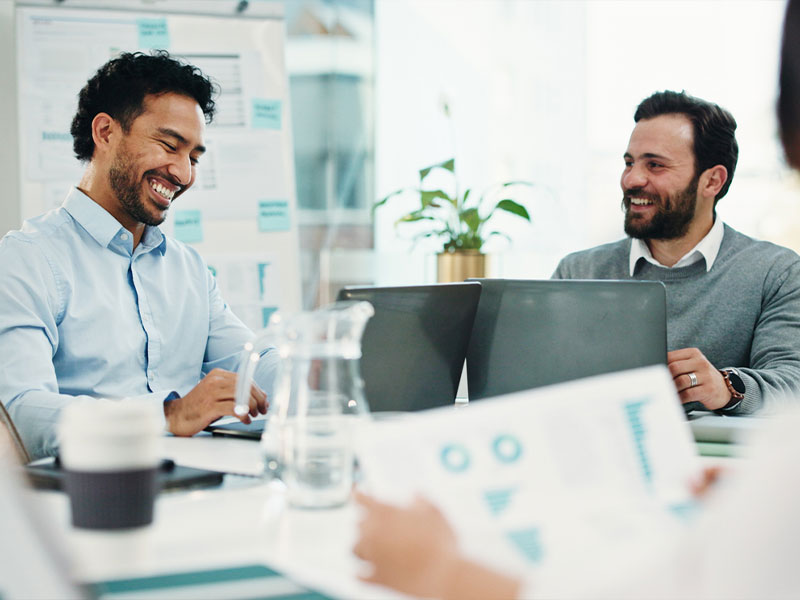 Two men smiling and working on laptops in a bright office meeting.