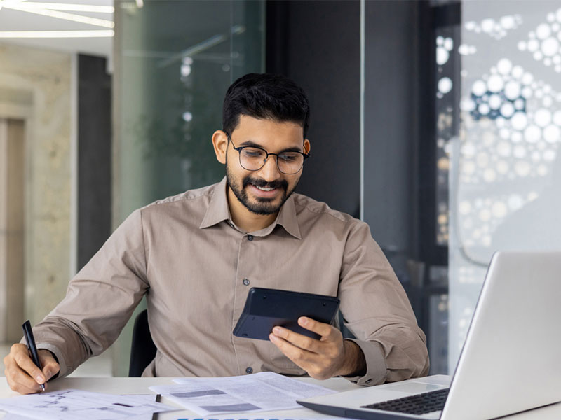 Man in an office reading from a tablet while writing, seated next to a laptop.