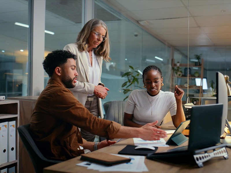 Three colleagues discussing a project at an office desk with documents and a laptop.