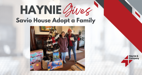 Haynie Gives event: two people stand with gifts for Savio House Adopt a Family near a decorated Christmas tree.