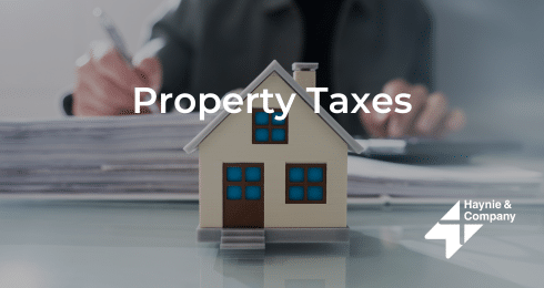 Toy house with Property Taxes text in front, person in background with documents, logo of Haynie.