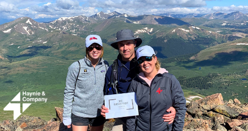 Three people in hiking gear on a mountain peak holding a sign, with vast scenic views in the background.