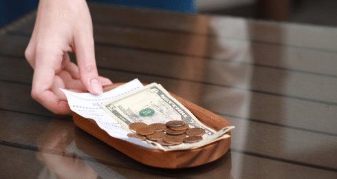 Hand placing cash and coins on a wooden tray with a receipt on a glass table.