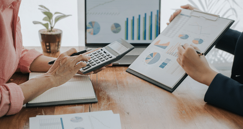Two people analyzing financial graphs with a calculator and clipboard on a wooden table.