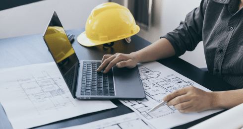 Engineer working with laptop and blueprints, safety helmet nearby.