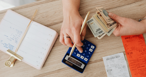 A person calculating finances with a calculator, cash, and receipts on a wooden desk.