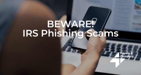Person holding phone with warning text about IRS phishing scams on screen.