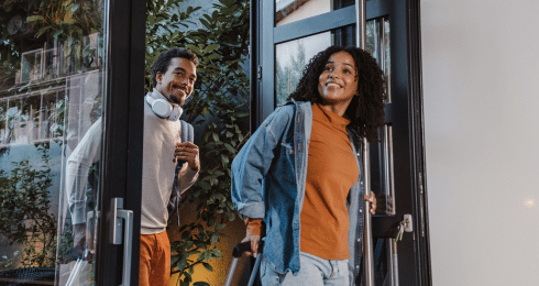 Two people smiling, entering a building with luggage, wearing casual clothes and backpacks near glass doors.