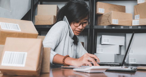 A woman multitasking with a phone and laptop among packages in a warehouse.