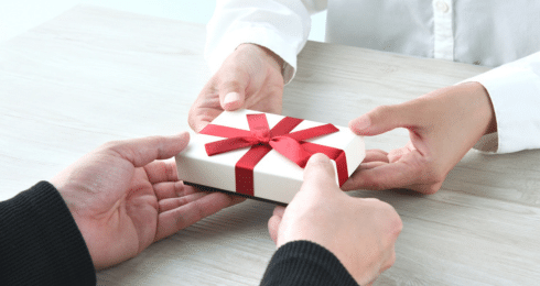 Hands exchanging a gift box wrapped with a red ribbon on a wooden table.
