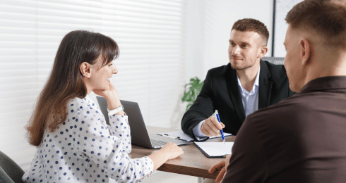 Three people in a business meeting, seated at a table with a laptop and documents.