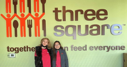 Two people standing in front of Three Square logo with green background.