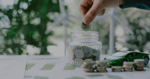 Hand placing coin in jar next to toy car, symbolizing financial savings and investment.