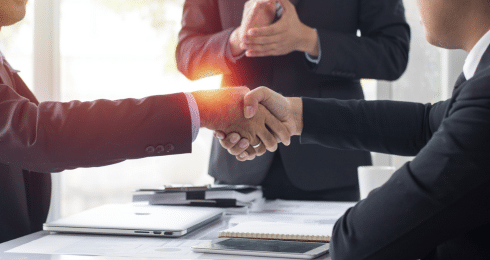 Business handshake in office setting with laptops and notepads on the table.