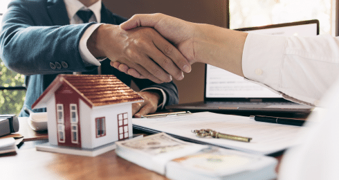 Real estate deal handshake over documents with house model on table.