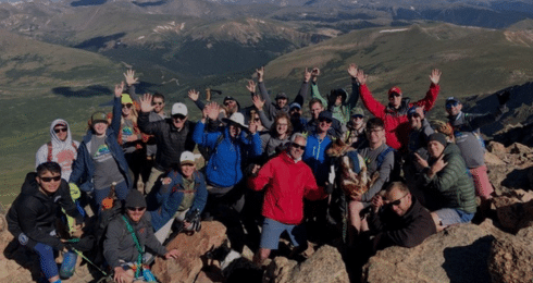 A group of people on a mountain summit celebrating with raised hands and scenic views in the background.