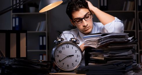 Man overwhelmed with paperwork, sitting at desk with clock, working late.