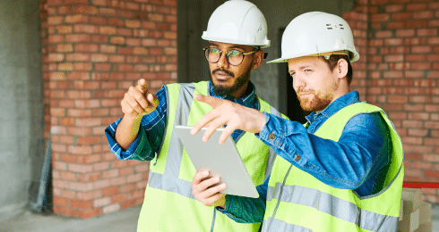 Two construction workers in safety vests and helmets reviewing a tablet on site.