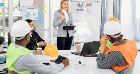 A woman leading a meeting with construction workers in hard hats at a table with plans and charts.