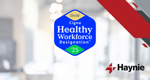 Cigna Healthy Workforce Designation Gold 2025 badge with Haynie logo and blurred office background.
