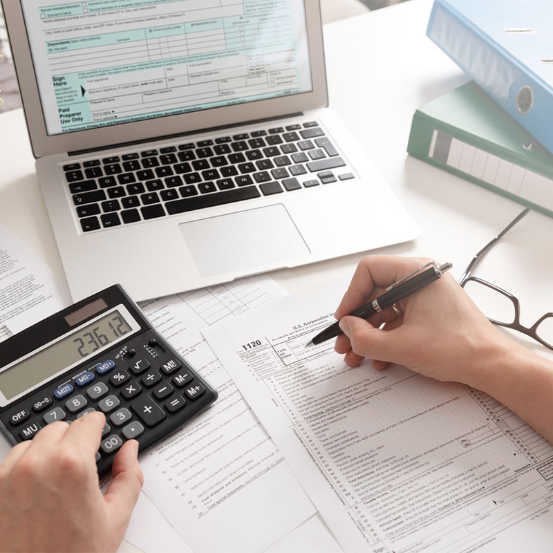 Person filling out tax forms with a calculator and laptop on a desk.