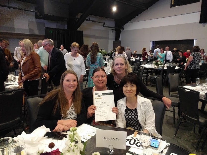 Group of four women at a corporate event, one holding a paper titled Up Up and Away, with others in the background.
