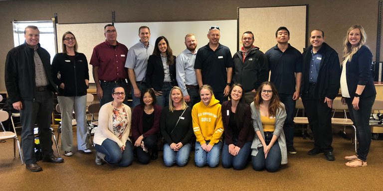 Group of people smiling in a classroom setting, some seated, some standing.