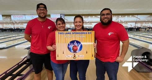 Four people holding a sponsor sign at a bowling alley, wearing matching red shirts.