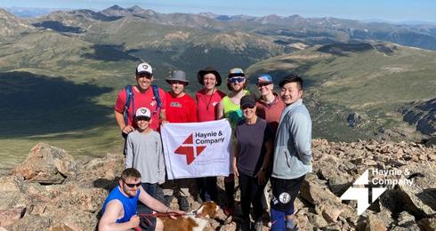 Group on mountain peak holding Haynie flag, scenic view in background.