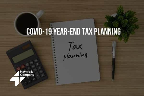 COVID-19 year-end tax planning notebook, calculator, coffee cup, plant, and pen on wooden desk.