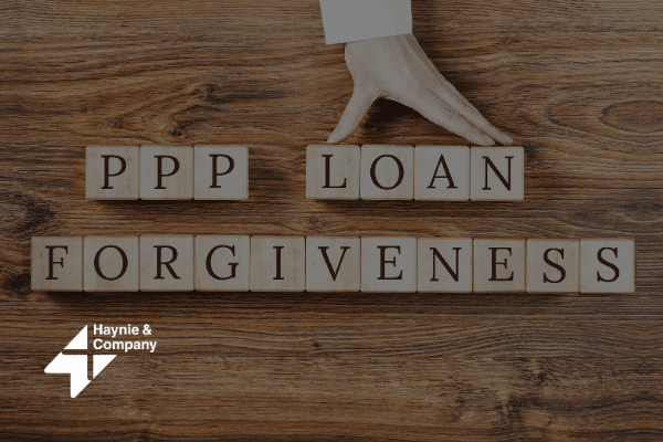 PPP Loan Forgiveness blocks on wooden table with a hand. Haynie logo.