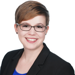 A person with short hair and glasses, wearing a black blazer and blue shirt, smiling at the camera.
