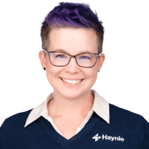 A person with glasses and purple hair, wearing a Haynie sweater, smiling in a professional portrait.