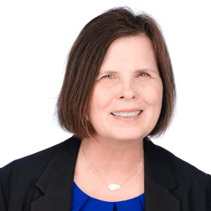 A smiling woman in a black blazer with a blue top, white background.