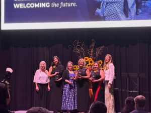 Women posing with awards in front of a stage backdrop reading Welcoming the Future, surrounded by sunflowers.