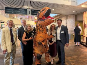 A group of people posing with a dinosaur costume at an indoor event.