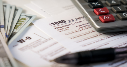 Tax forms, calculator, pen, and money on a table.