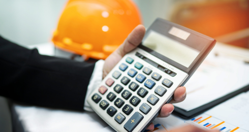 Person in suit holding calculator near construction helmet and charts.