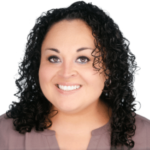 A person smiling with curly hair, wearing a gray top against a white background.
