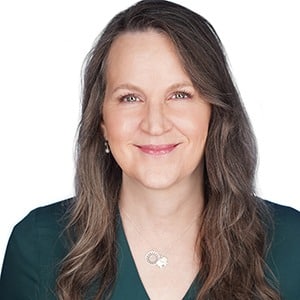 A smiling woman with long brown hair wearing a dark green top against a white background.