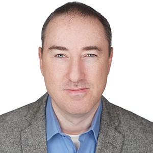 A person wearing a blue shirt and gray blazer smiles against a white background.